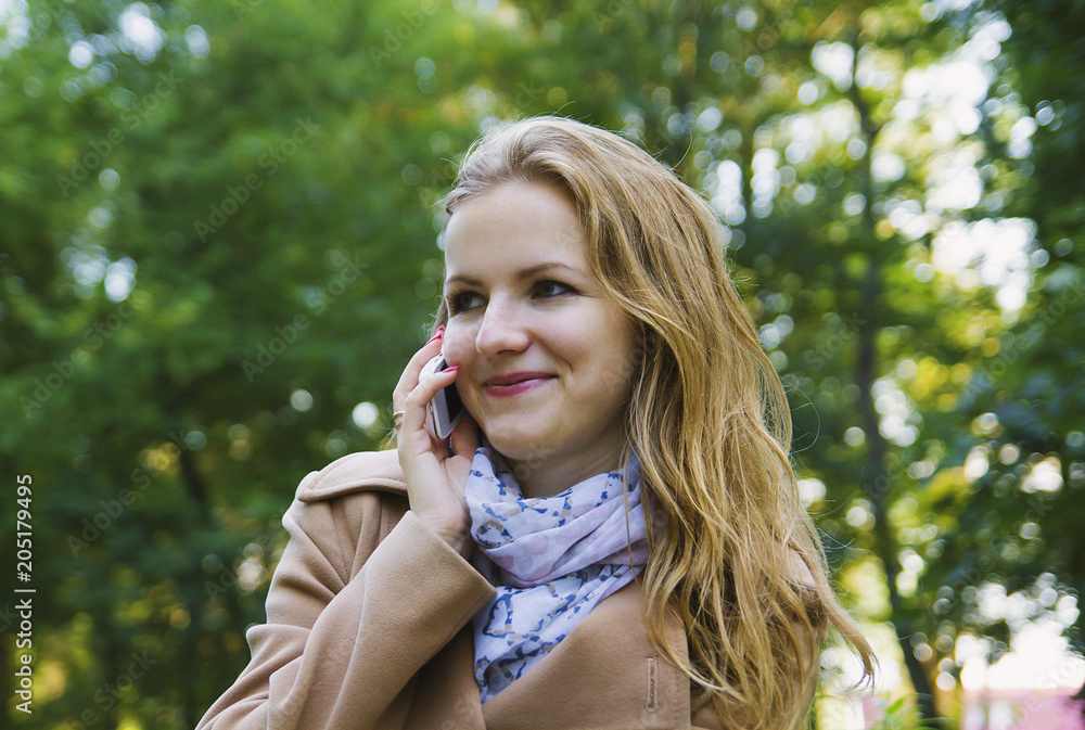 Beautiful girl with fair hair is talking on phone in green park. Young woman in scarf and beige coat. Cute autumn style photo. Beautiful girl with fair hair is talking on phone in green park. Young woman in scarf and beige coat. Cute autumn style photo.