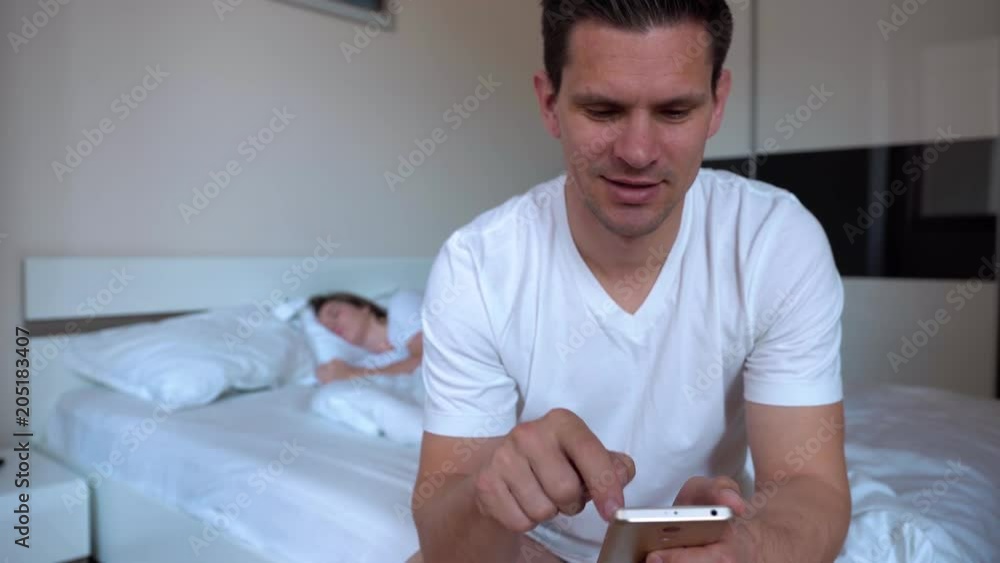 Happy Young Man Playing With Smartphone On The Background Of Sleeping Wife Before Night Sleep Happy Young Man Playing With Smartphone On The Background Of Sleeping Wife Before Night Sleep