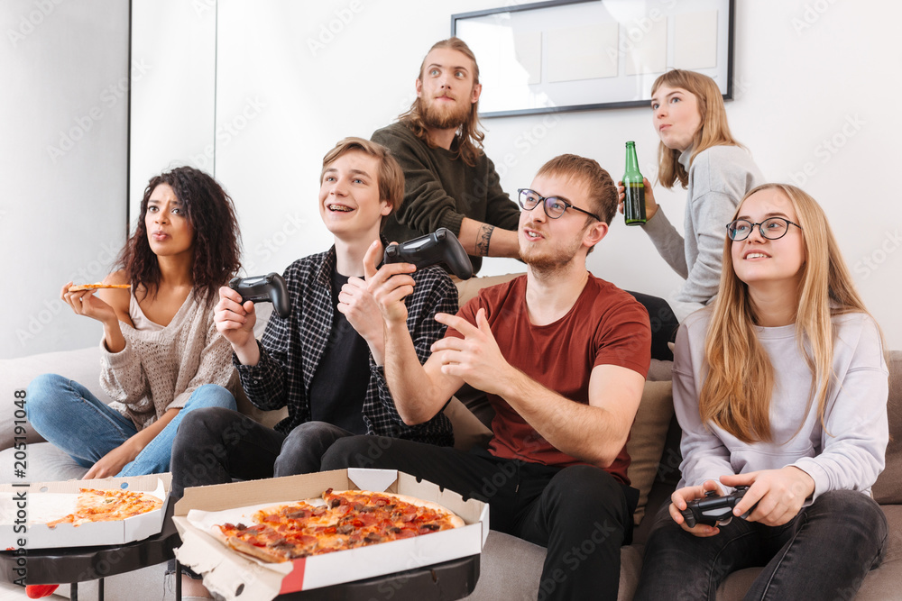 Group of friends sitting on sofa and spending time together while playing video games,eating pizza and drinking beer at home. Portrait of young people showing different emotions isolated Group of friends sitting on sofa and spending time together while playing video games,eating pizza and drinking beer at home. Portrait of young people showing different emotions isolated