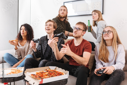 Group of friends sitting on sofa and spending time together while playing video games eating pizza and drinking beer at home. Portrait of young people showing different emotions isolated