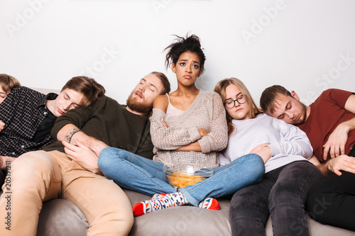 Portrait of young upset lady sitting on sofa with chips and sadly looking aside while her friends sleeping near at home