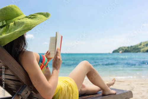 Side view of a young beautiful woman reading a book while sitting on a wooden lounge chair at the beach in a sunny day during summer vacation in Flores Island, Indonesia