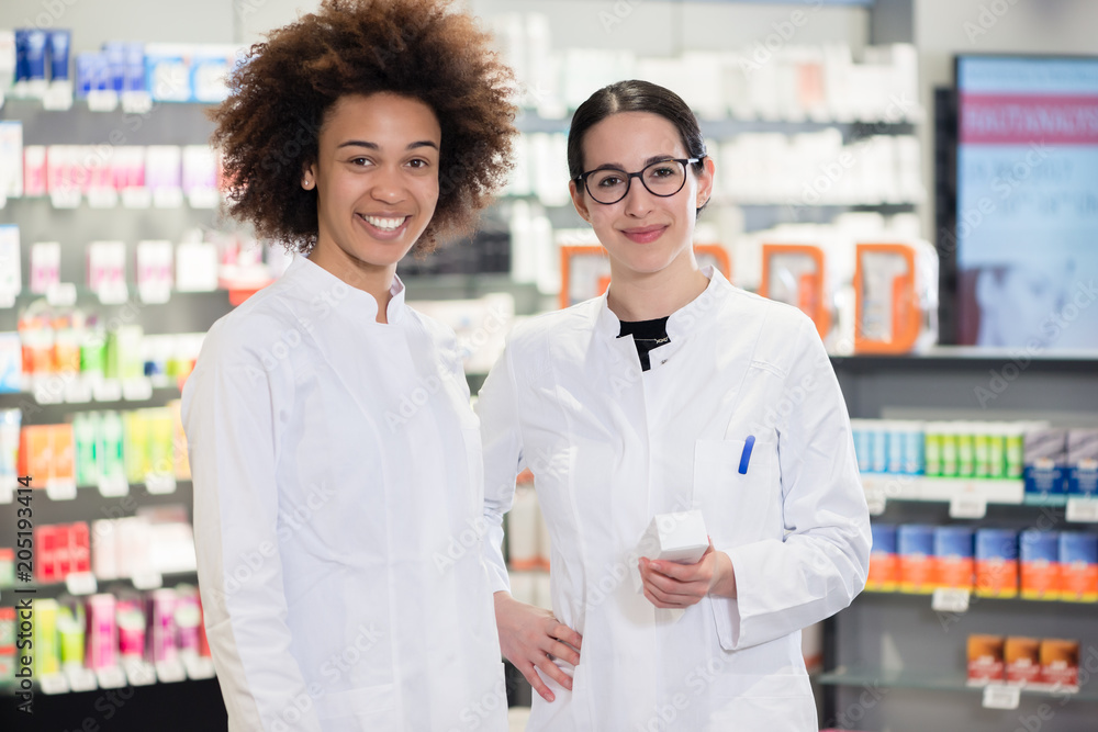 Portrait of two female pharmacists wearing white lab coats, while smiling with confidence at work in the interior of a modern drugstore with various products for sale