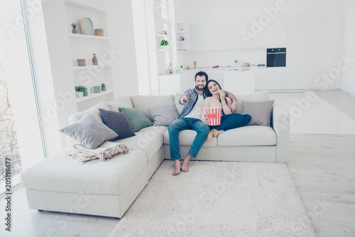 Full size portrait of stylish positive couple sitting in modern white apartment using console looking comedy funny channel program film on television hugging embracing eating snack © deagreez