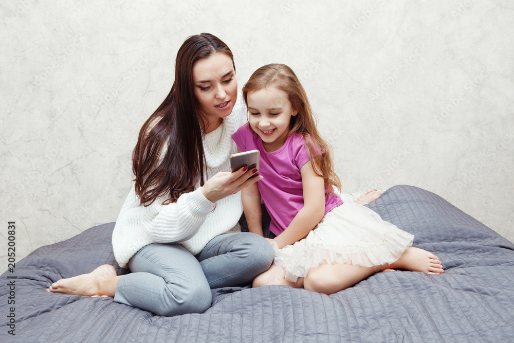 Young woman and girl are looking together at the mobile phone screen. They are sitting at a table on which there is a laptop. It's mom and daughter. Young woman and girl are looking together at the mobile phone screen. They are sitting at a table on which there is a laptop. It's mom and daughter.