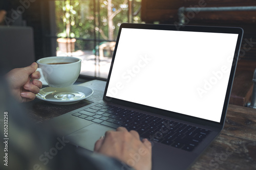Mockup image of a woman using laptop with blank white desktop screen while drinking hot coffee in cafe