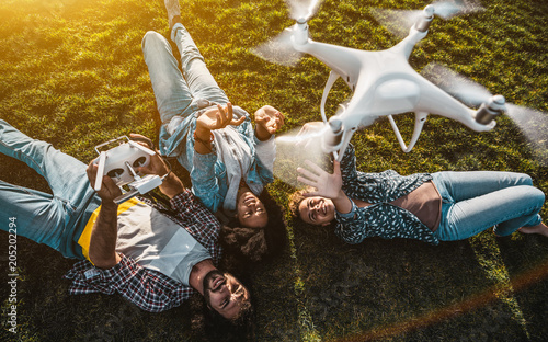 The group of happy curly people: Caucasian guy, an African-American girl, and a Caucasian girl are laying on the summer meadow while recording vlog video using flying drone operated by the man