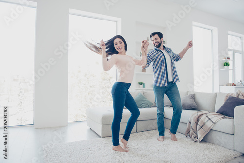 Portrait of active cheerful couple in casual outfits dancing singing indoor, in modern open space living room enjoying time together