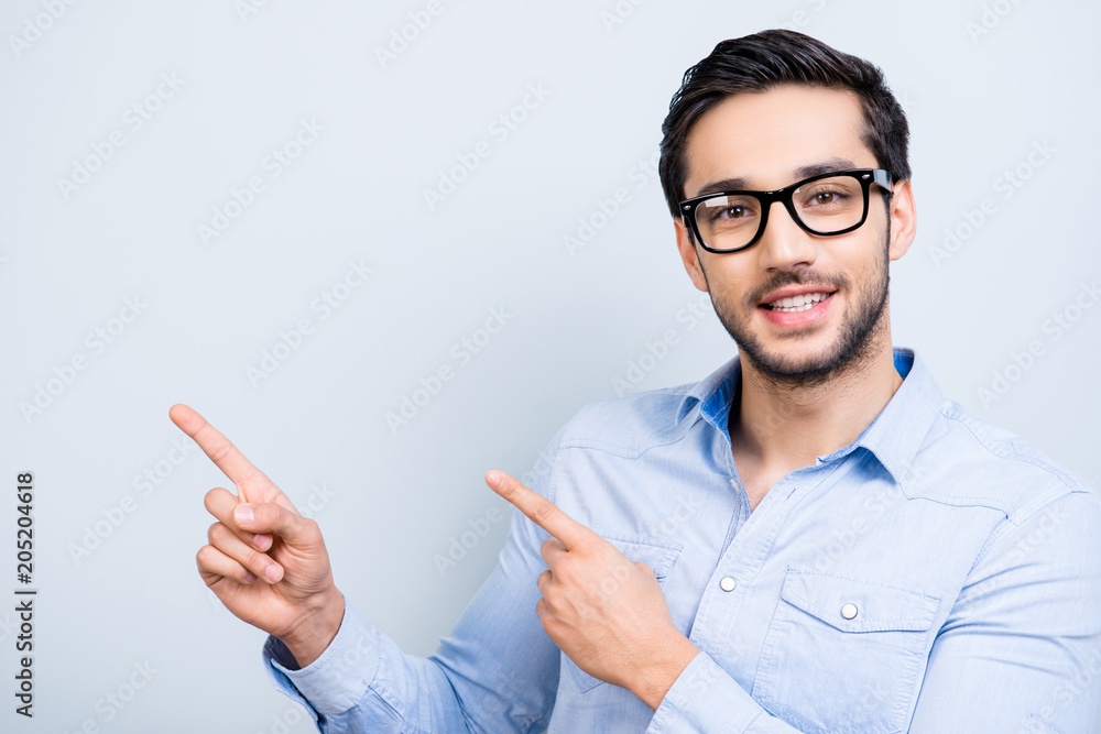Portrait with copyspace, cute smiling man with black hair in glasses indicate showing empty place with two forefingers looking at camera isolated on grey background, direction way decision concept Portrait with copyspace, cute smiling man with black hair in glasses indicate showing empty place with two forefingers looking at camera isolated on grey background, direction way decision concept