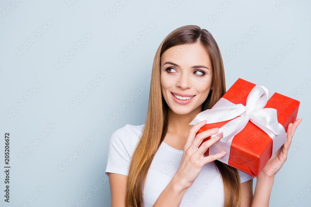 Portrait with copyspace empty place of nice gorgeous girl holding gift box in red package with white ribbon near ear want to know what inside isolated on grey background Portrait with copyspace empty place of nice gorgeous girl holding gift box in red package with white ribbon near ear want to know what inside isolated on grey background