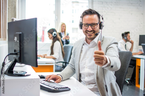 Cheerful business man with headset showing thumb up while working on computer in call center.
