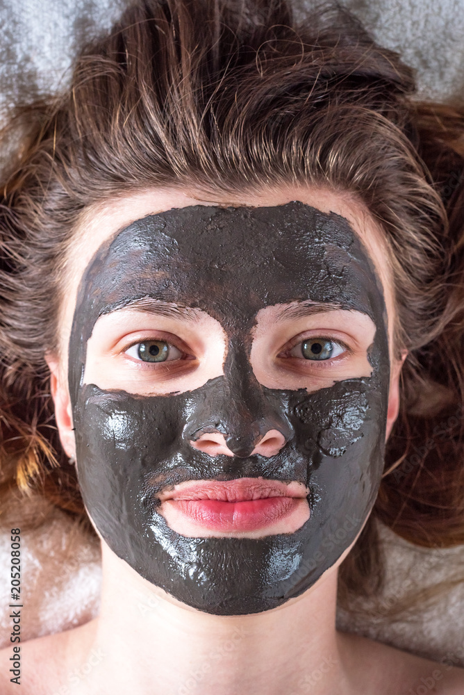 Fototapeta premium A young woman with her hair and a cosmetic mask of black clay on her face - cosmetic procedures close-up