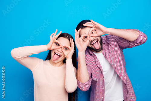 Portrait of cheerful joyful couple in casual outfit making binoculars with fingers gesturing tongue out isolated on blue background