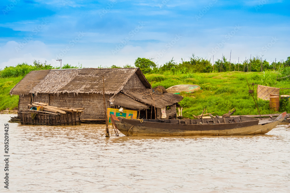 A floating house made of wood and thatch with a tied up wooden boat along the Siem Reap River in Cambodia. A floating house made of wood and thatch with a tied up wooden boat along the Siem Reap River in Cambodia.