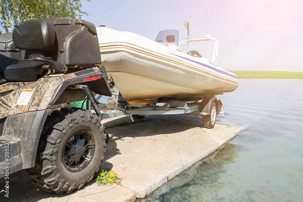 Obraz premium Transportation of inflatable boat on trailer. ATV quadbike moves ship to lake or river shore for launching. Beginning of water navigation and fishing season