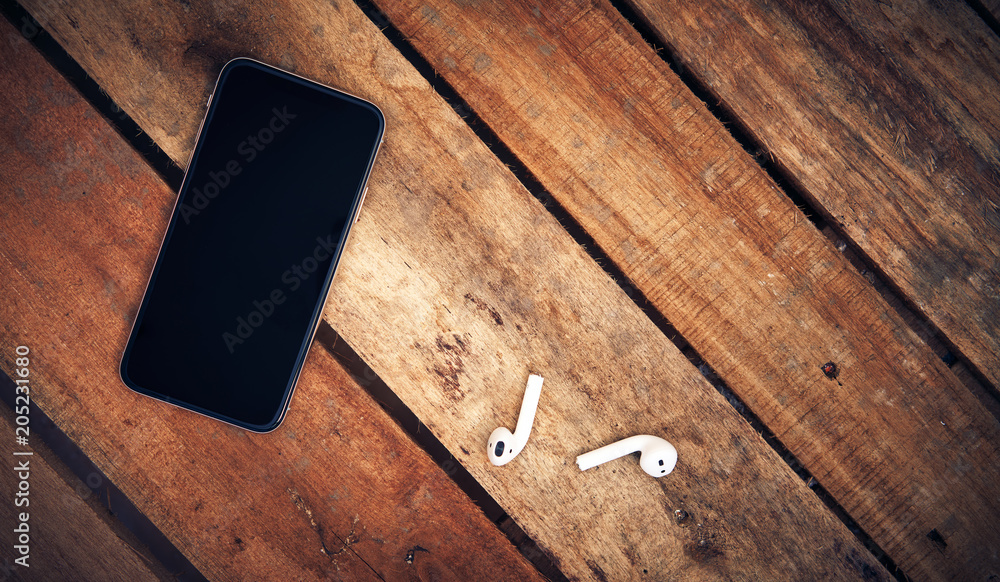 A top view of a set of wireless earbud headphones and mobile phone blank template lying on a rustic wooden table. Styling and grain effect added to image.