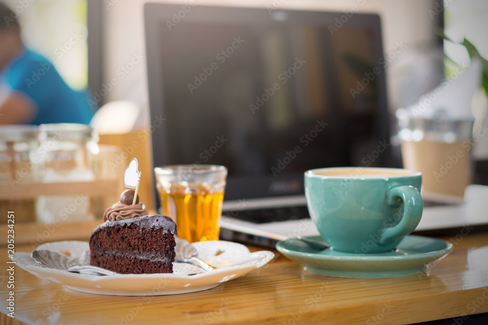 Chocolate cake and coffee cup are on the table in the cafe with a notebook as the background.