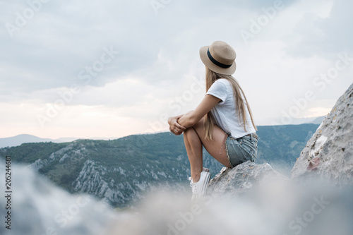 Young traveling woman wearing hat sitting high on the top of the mountain, evening with sunset. Montenegro