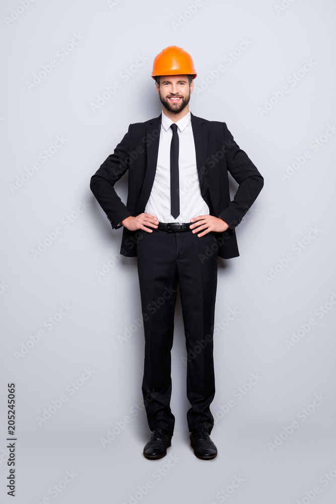 Full length size portrait of attractive cheerful joyful investor in orange hard hat and black tuxedo with tie, looking at camera, isolated on grey background, holding two arms on waist Full length size portrait of attractive cheerful joyful investor in orange hard hat and black tuxedo with tie, looking at camera, isolated on grey background, holding two arms on waist