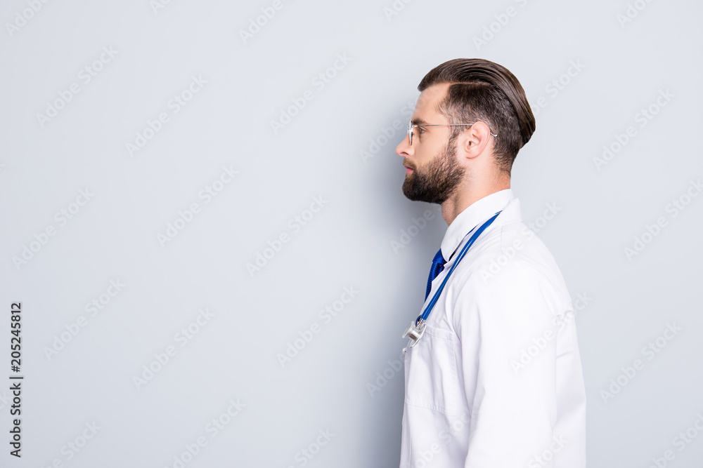 Profile portrait with copy space, empty place for advertisement of attractive stylish doc with stubble in white lab coat and stethoscope on his neck, isolated on grey background Profile portrait with copy space, empty place for advertisement of attractive stylish doc with stubble in white lab coat and stethoscope on his neck, isolated on grey background