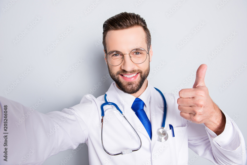 Self portrait of positive trendy doc in white outfit with tie and bristle having stethoscope on his neck shooting selfie showing thumb up, like sign with finger, hand over grey background Self portrait of positive trendy doc in white outfit with tie and bristle having stethoscope on his neck shooting selfie showing thumb up, like sign with finger, hand over grey background