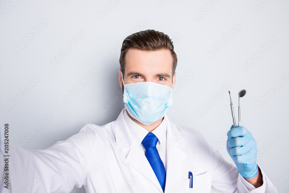 Self portrait of successful cheerful dentist in white lab coat, blue tie, shooting selfie on front camera, holding tools, having leisure, fun, video call, isolated on grey background Self portrait of successful cheerful dentist in white lab coat, blue tie, shooting selfie on front camera, holding tools, having leisure, fun, video call, isolated on grey background