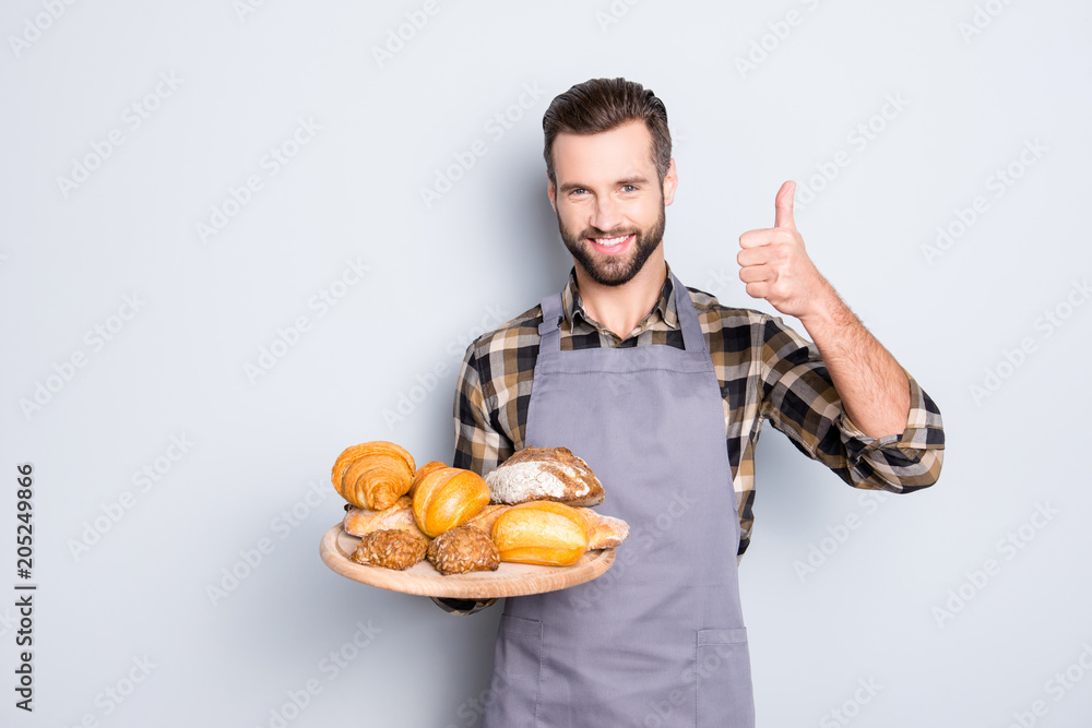 Portrait of positive attractive baker in uniform with stubble holding tray with bakery products in hand, showing thumb up with finger, isolated on grey background Portrait of positive attractive baker in uniform with stubble holding tray with bakery products in hand, showing thumb up with finger, isolated on grey background