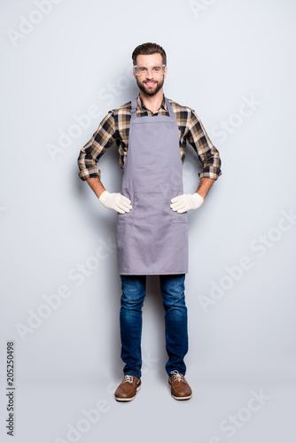 Full size body portrait of attractive cheerful carpenter with hairstyle in safety glasses, jeans, looking at camera holding his two arms on waist isolated on grey background photo