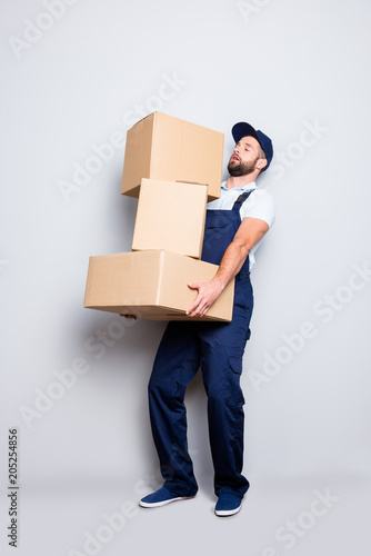 Full size body portrait of busy attractive deliver in blue uniform with stubble, having three heavy big boxes in arms, isolated on grey background, trying to withhold