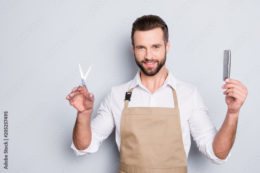 Portrait of cheerful, joyful barber with stubble in shirt having scissors, tools, equipments, comb in hands looking at camera isolated on grey background Portrait of cheerful, joyful barber with stubble in shirt having scissors, tools, equipments, comb in hands looking at camera isolated on grey background