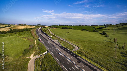 Italia, Maggio 2018 - Vista aerea dell'autostrada con automobili e camion che attraversa le colline con i campi di grano non ancora maturo