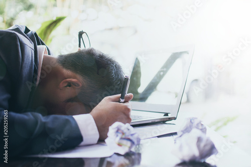 Fotografija Stressed businessman in dark suit sitting at office desk full with books and pap