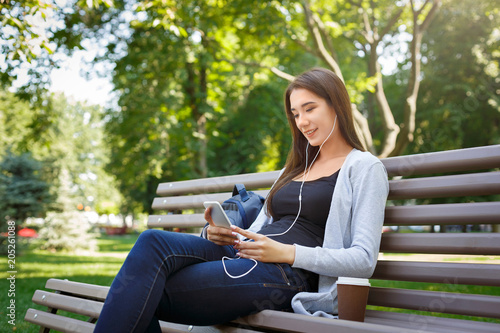 Smiling young female IT student in headphones. Video calling and drinking coffee in the park