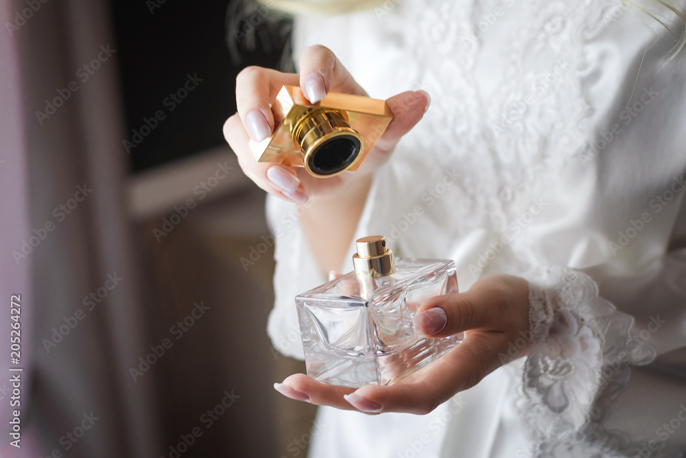 Close up shoot of a luxury fragrant perfume flacon in manicured hands of elegant bride wearing white dressing gown - obrazy, fototapety, plakaty 