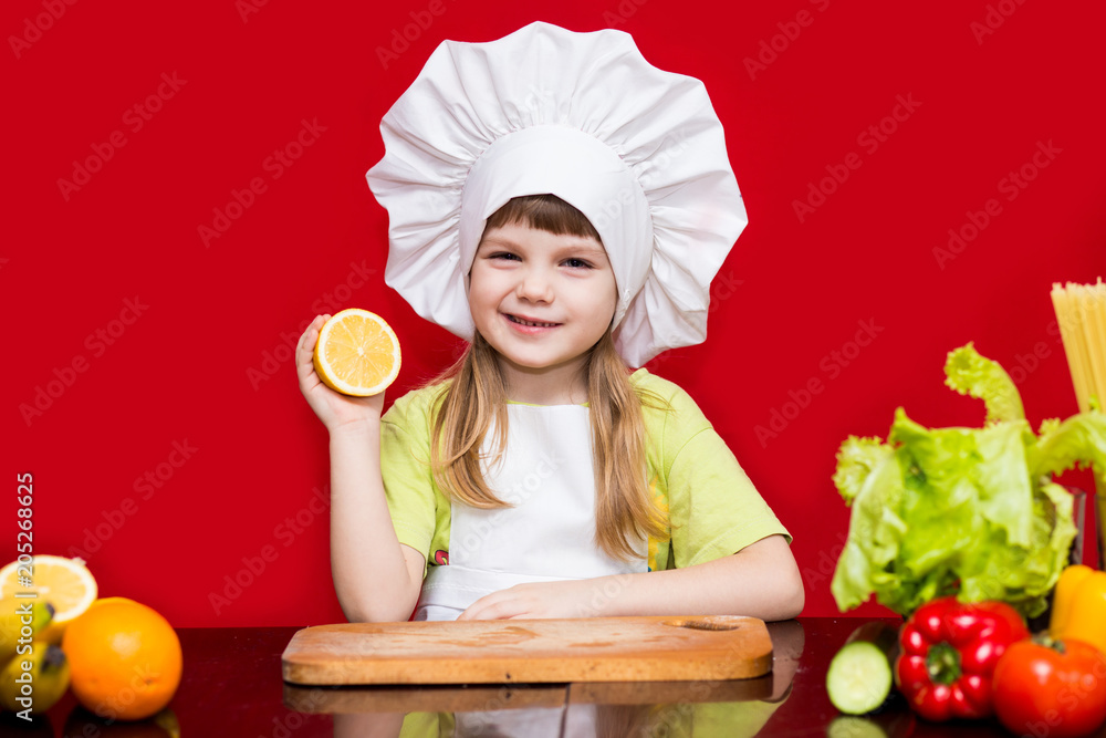 Happy little girl in chef uniform cuts fruit in kitchen. Kid chef. Cooking Process Concept Happy little girl in chef uniform cuts fruit in kitchen. Kid chef. Cooking Process Concept
