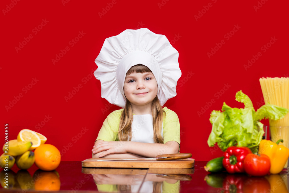 Happy little girl in chef uniform cuts vegetables in kitchen. Kid chef. Vegetarian. Cooking Process Concept Happy little girl in chef uniform cuts vegetables in kitchen. Kid chef. Vegetarian. Cooking Process Concept