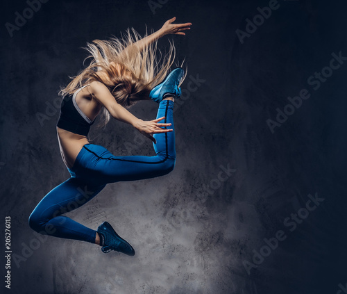 Fototapeta Naklejka Na Ścianę i Meble -  Young blonde ballerina in sportswear dances and jumps in a studio. Isolated on a dark background.