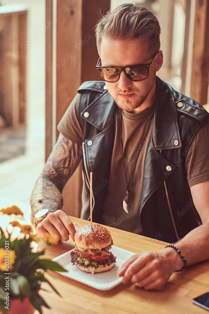 Handsome hipster with a stylish haircut and beard sits at a table, decided to dine at a roadside cafe, eating a hamburger.