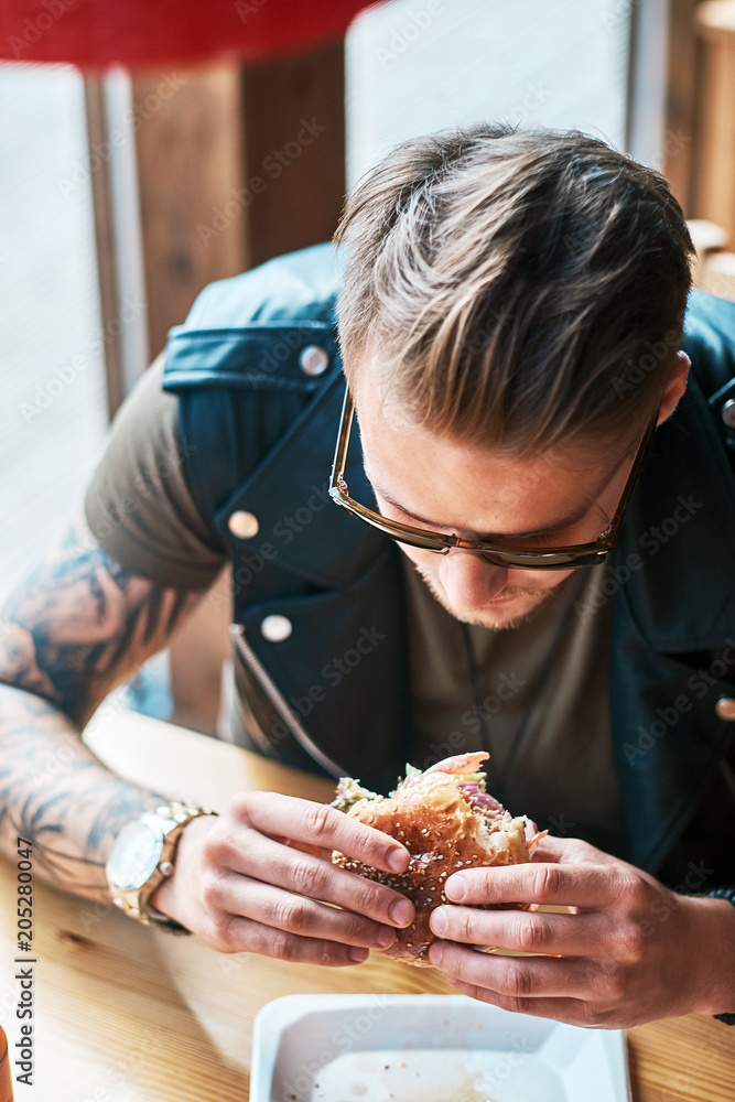 Portrait of a hungry hipster guy with a stylish haircut and beard sits at a table, decided to dine at a roadside cafe, eating a hamburger. Portrait of a hungry hipster guy with a stylish haircut and beard sits at a table, decided to dine at a roadside cafe, eating a hamburger.
