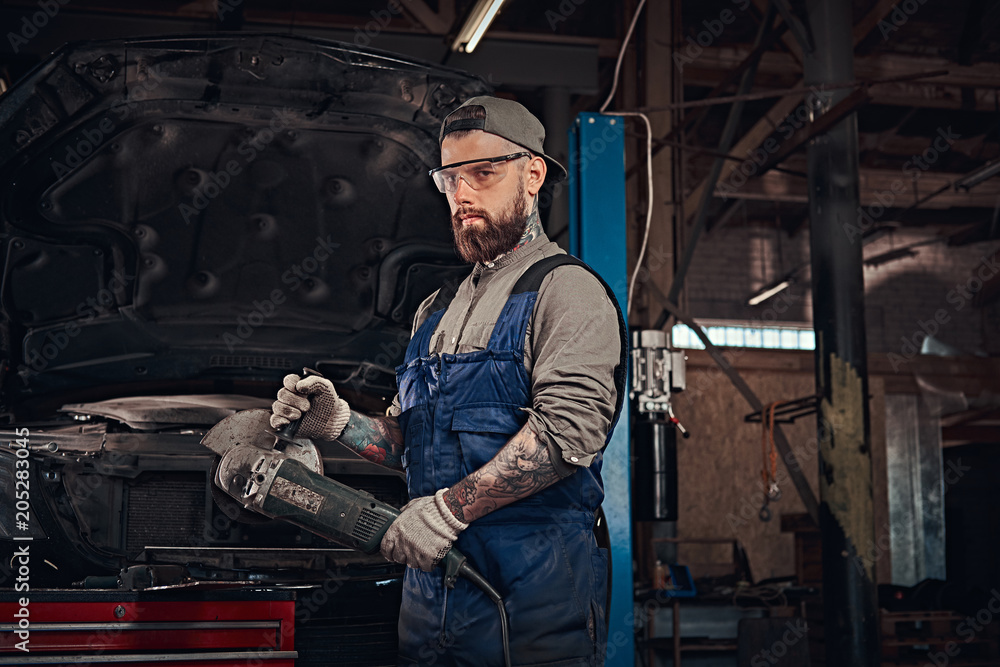 Brutal auto mechanic in a uniform and safety glasses working with an angle grinder while standing against a broken car in repair garage.