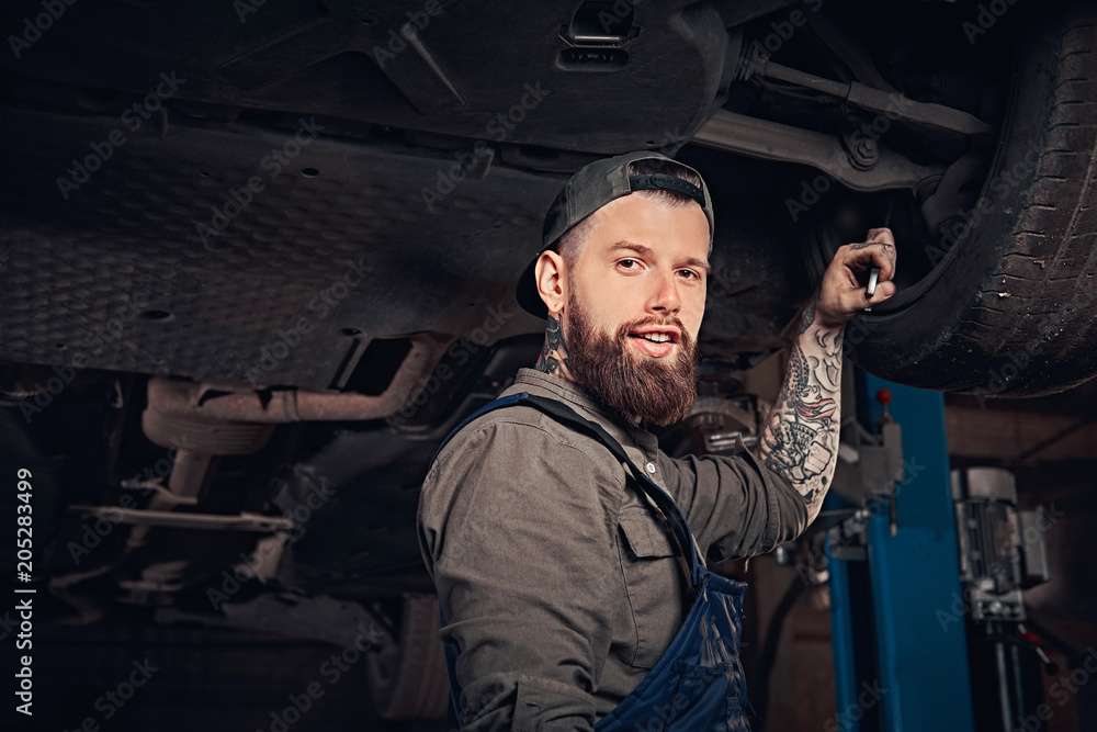 Bearded auto mechanic in a uniform repair the car's suspension with a wrench while standing under lifting car in repair garage.