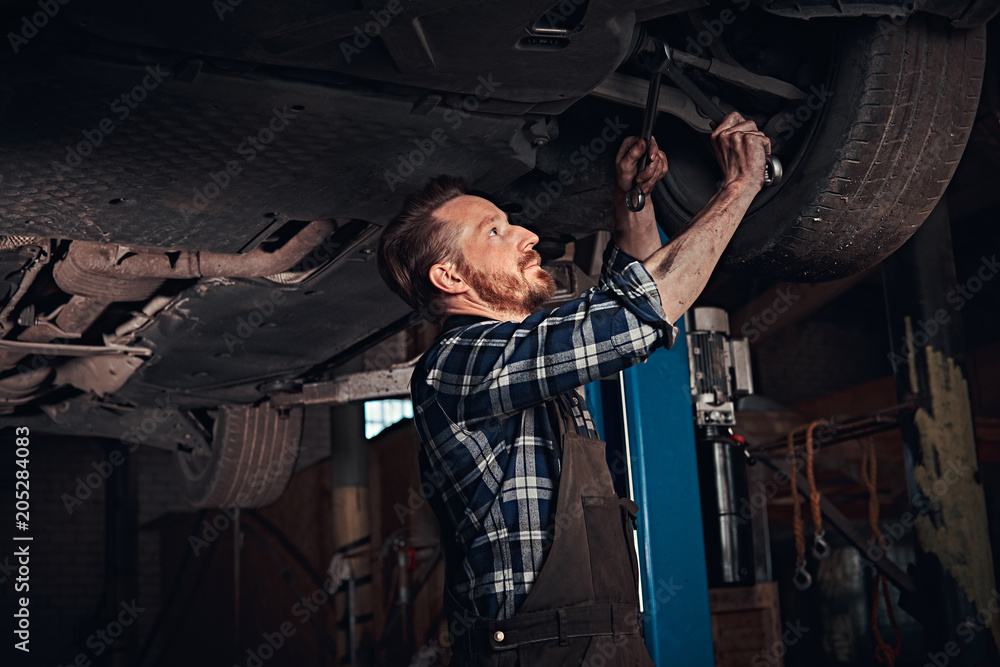 Bearded auto mechanic in a uniform repair the car's suspension with a wrench while standing under lifting car in repair garage.