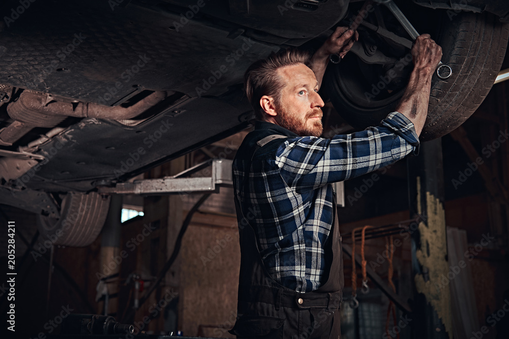 Bearded auto mechanic in a uniform repair the car's suspension with a wrench while standing under lifting car in repair garage. Bearded auto mechanic in a uniform repair the car's suspension with a wrench while standing under lifting car in repair garage.