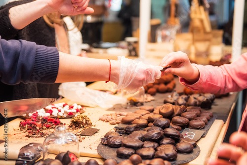 Display with assortment of dark and brown chocolate candies with different fillings. Seller's hand picks some slice for buyer testing. Tasty and delicious desserts. Selective focus.