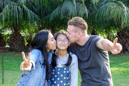 Family in park portrait. White blond man, chinese woman, chinese girl standing in park together and kissing. Family fun concept.