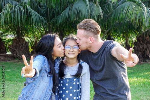 Family in park portrait. White blond man, chinese woman, chinese girl standing in park together and kissing. Family fun concept.