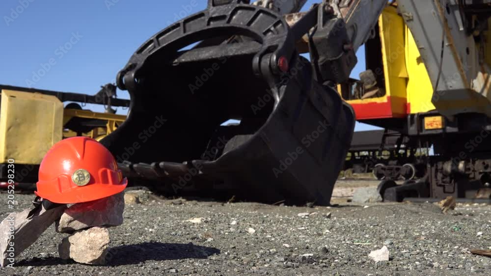 The golden bitcoin stands on an orange helmet against the background of a large bucket of a mining excavator. The golden bitcoin stands on an orange helmet against the background of a large bucket of a mining excavator.