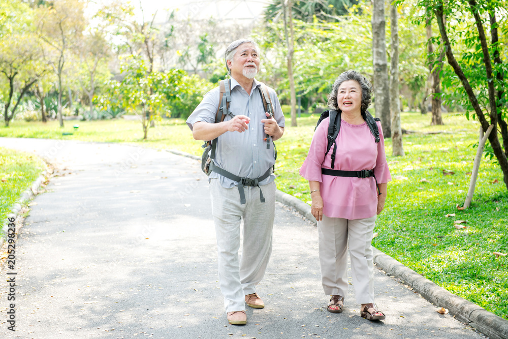 Fototapeta premium Senior couple in park walking with backpack. Chinese old couple in park, relaxing, smiling. With copy space.