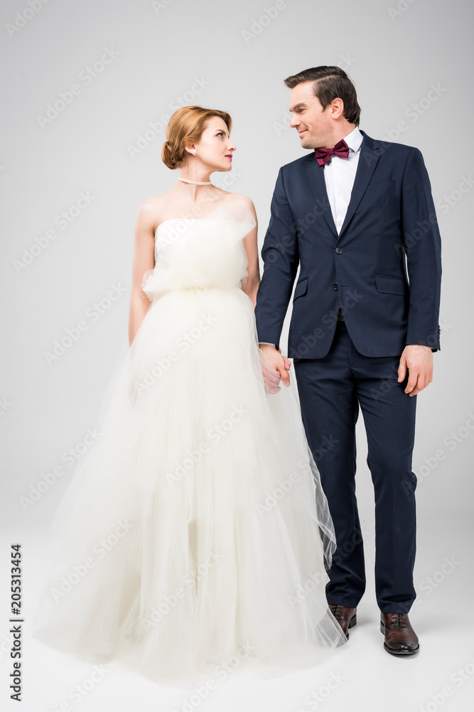groom in tuxedo and bride in wedding dress holding hands, isolated on grey