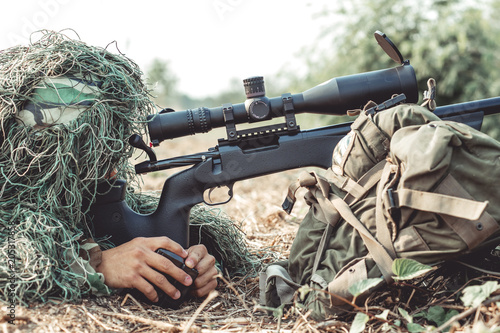 Sniper in urban combat training. Full ghillie camo suit and a high power sniper rifle. In urban combat training ground. Ready his weapon.